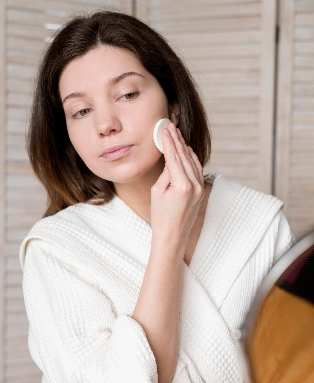 Femme en peignoir blanc appliquant un soin du visage avec un coton devant un miroir.
