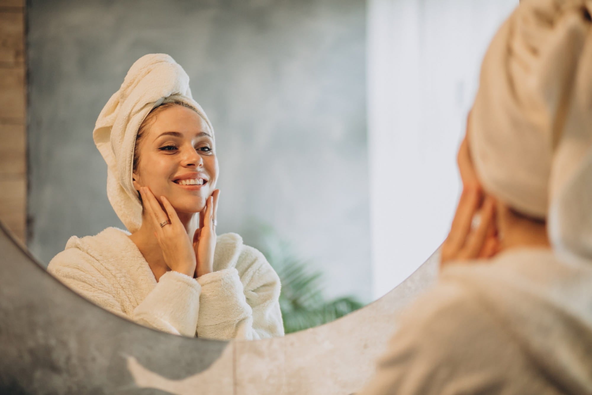Femme en peignoir blanc souriant face au miroir de sa salle de bain pendant son rituel de soin visage